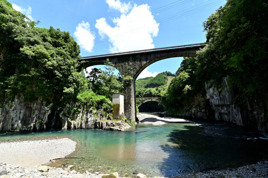 川の両岸を結ぶ出会橋を下から見上げた景色。崖の間に並ぶ二つのアーチと、青空・緑が調和する風景