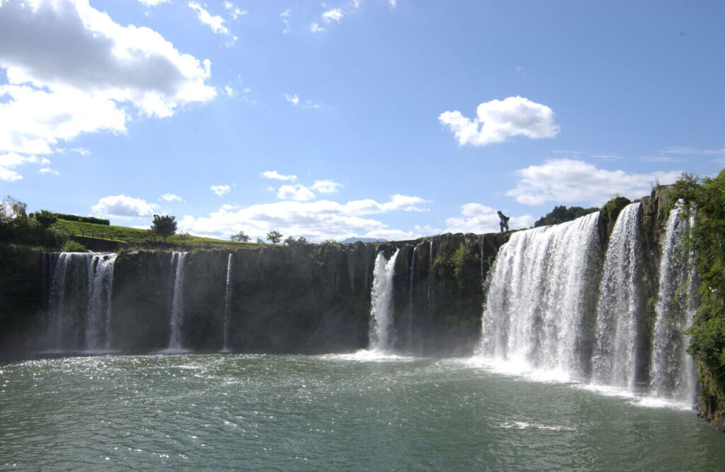 原尻の滝（はらじりのたき）を右方向から望む風景。青空と白い雲を背景に、阿蘇火山の噴火で生まれた大分県豊後大野市の壮大な滝が広がる