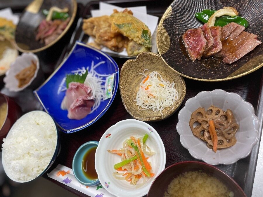 民宿いこいの夕食膳。和牛ステーキ、地魚の刺身、天ぷら、煮物、白米、味噌汁が並ぶグレードアップ膳の一例
