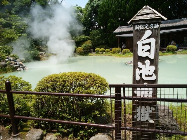 白池地獄の立て看板の奥に、乳白色の湯面と湯気が広がる風景