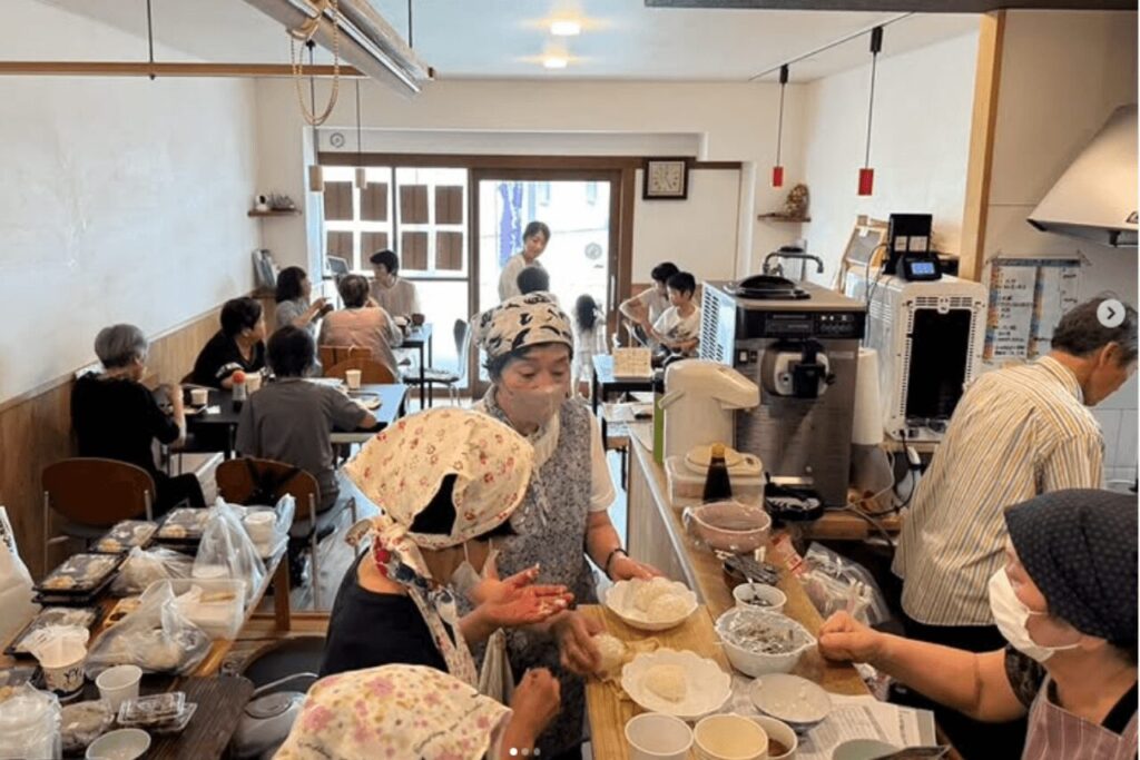 大分市子ども食堂リトルダイナーでNPOボランティアが料理を準備し、地域の人々がテーブルで交流する様子