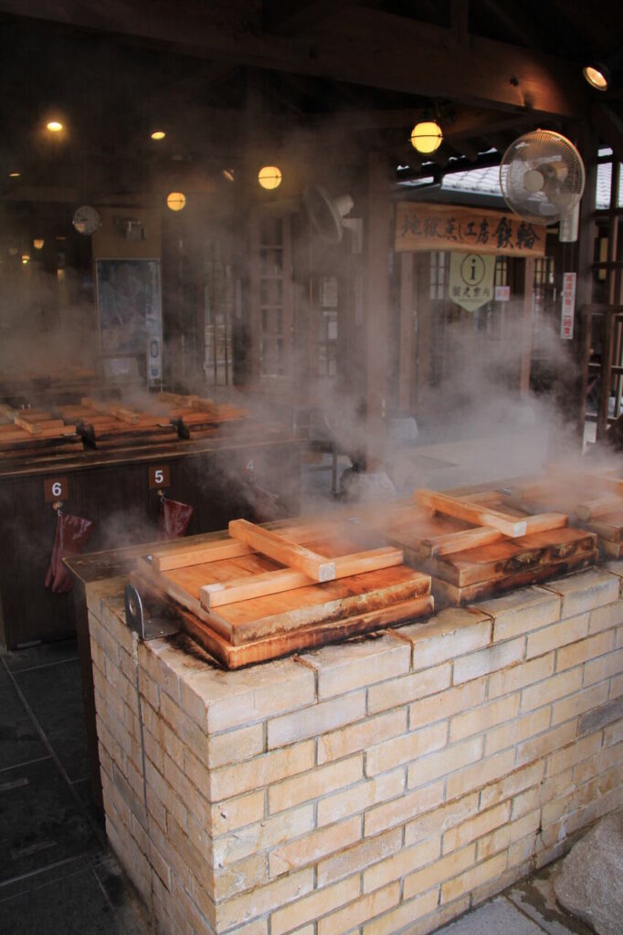 別府・地獄蒸し工房で木の釜が並び、湯気が立ち上る地獄蒸しの様子
