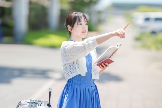 観光地を指さす旅行用バッグと旅行本を持つ女性