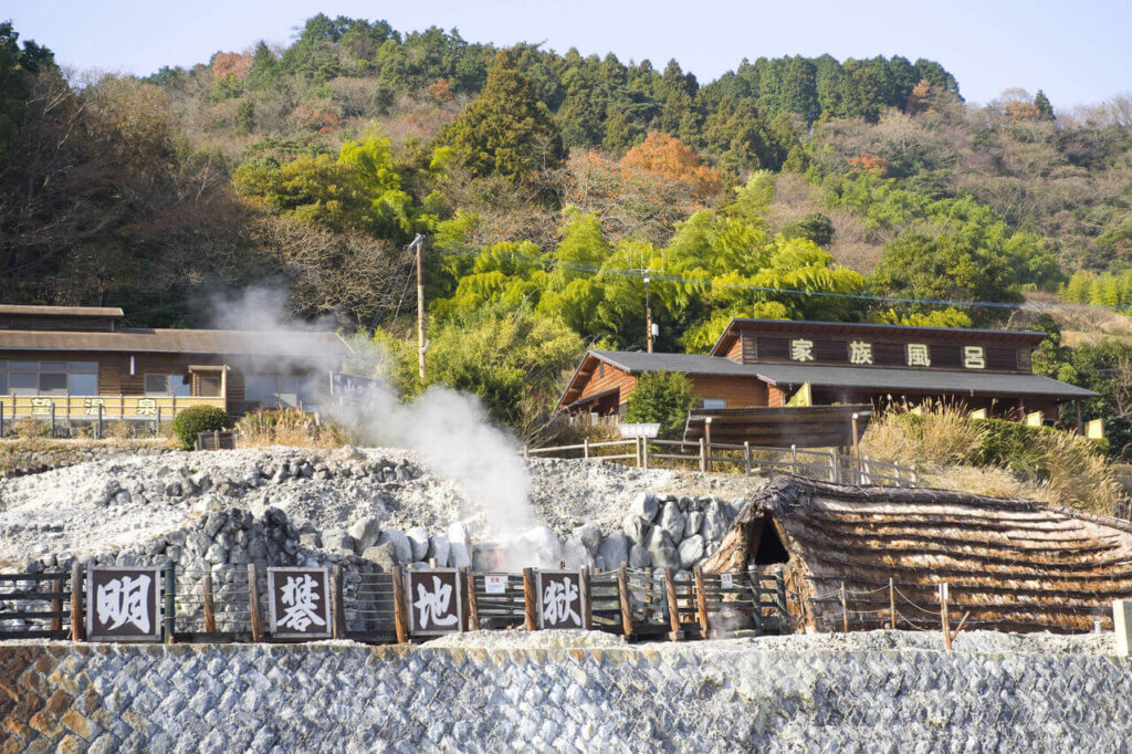 明礬地獄の看板と、遠くに見える山の湯を見上げる風景