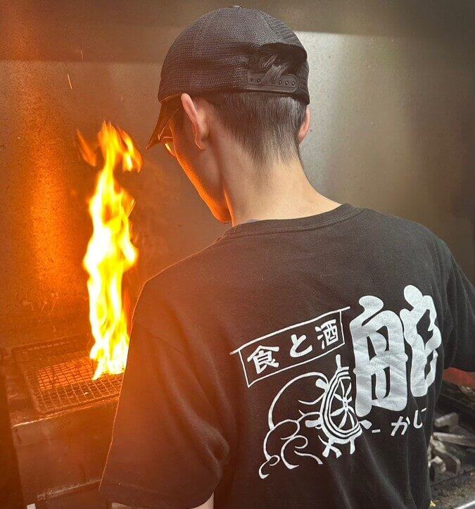 大分県の食と酒 舵の店内の様子。調理場所の店員の後ろ姿。地鶏を焼いていて火柱が経っている