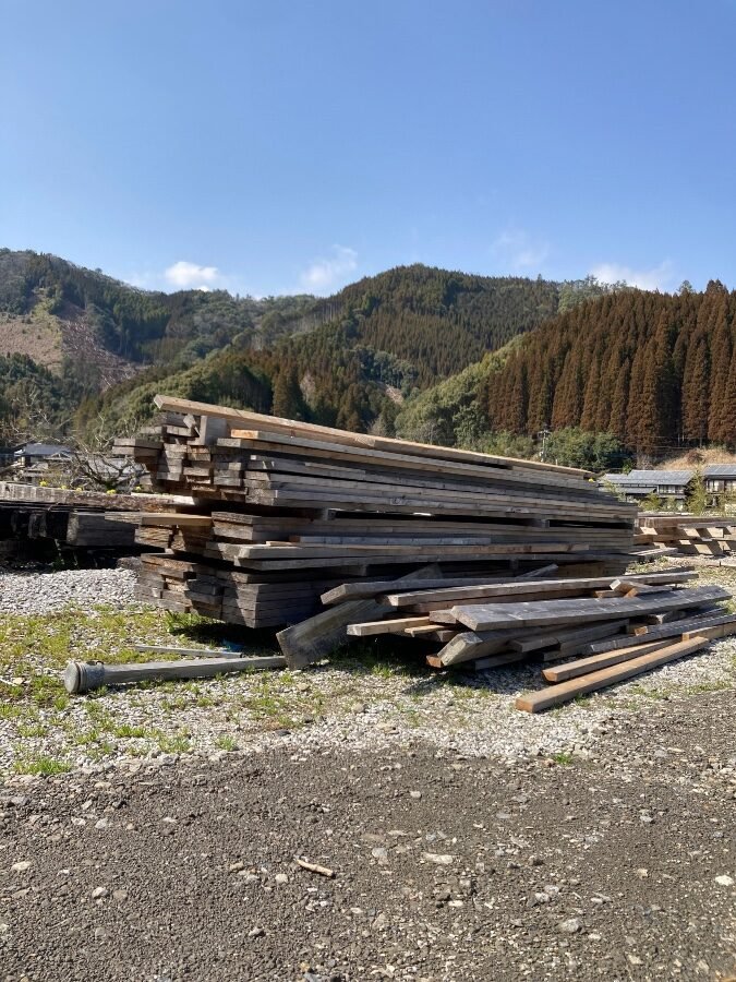 直川の地元企業から提供された廃材の角材が積み上げられた様子。山の景色を背景にした屋外の廃材置き場
