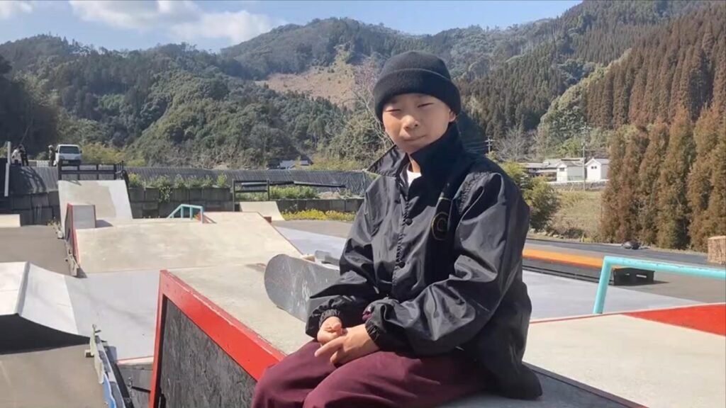 スケートボードを手にしたスケーターがコース上に座る様子。背後にはRefine Skate Parkのランプやコース全景、山の緑が広がる