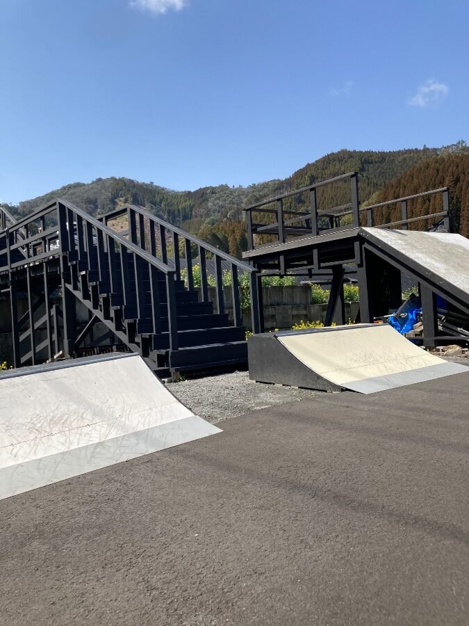 Refine Skate Parkのミニランプと黒い木製の階段型ストラクチャー。青空と直川の山並みを背景にした手作りコースの全景