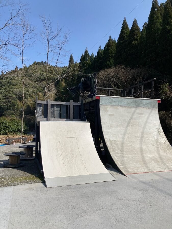 2連のハーフパイプ（ミニランプ）。奥のデッキからスケーターが滑り出す瞬間。背後は杉林と青空
