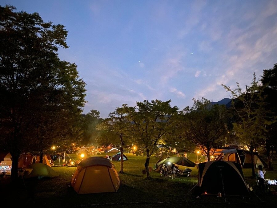 夕暮れから夜へと変わる空の下、多数のテントが灯りをともすフリーサイトの幻想的な夜景
